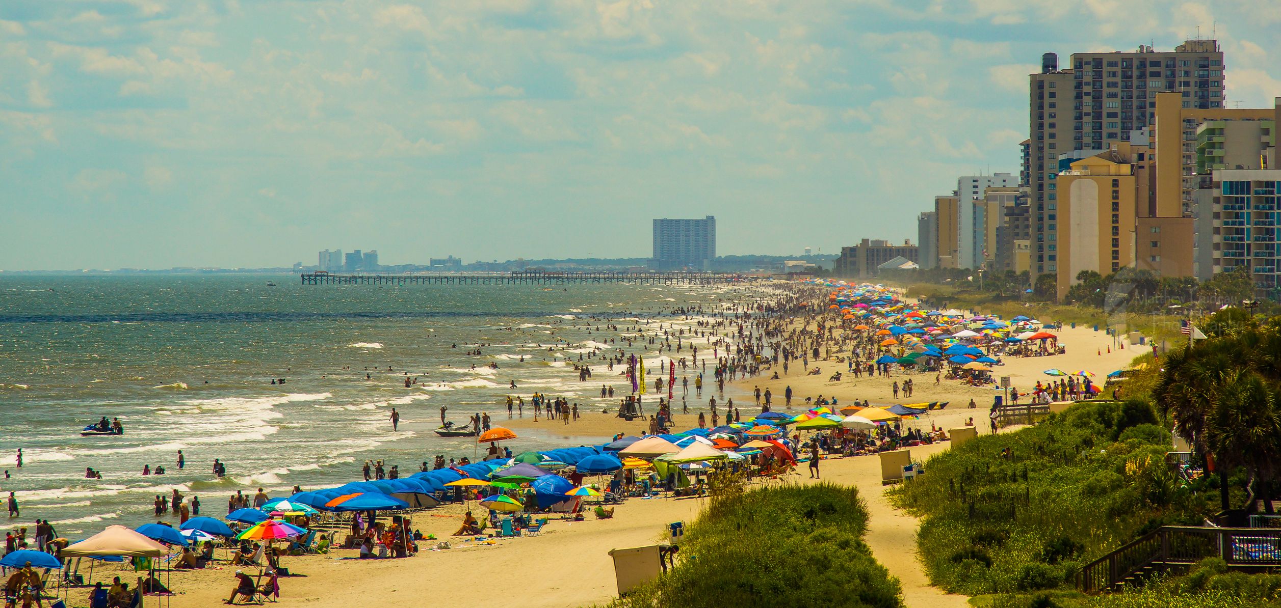 A busy Myrtle Beach oceanfront with colorful beach umbrellas, hotels, and a fishing pier in the distance; beach context near the Boardwalk venue, not the event.