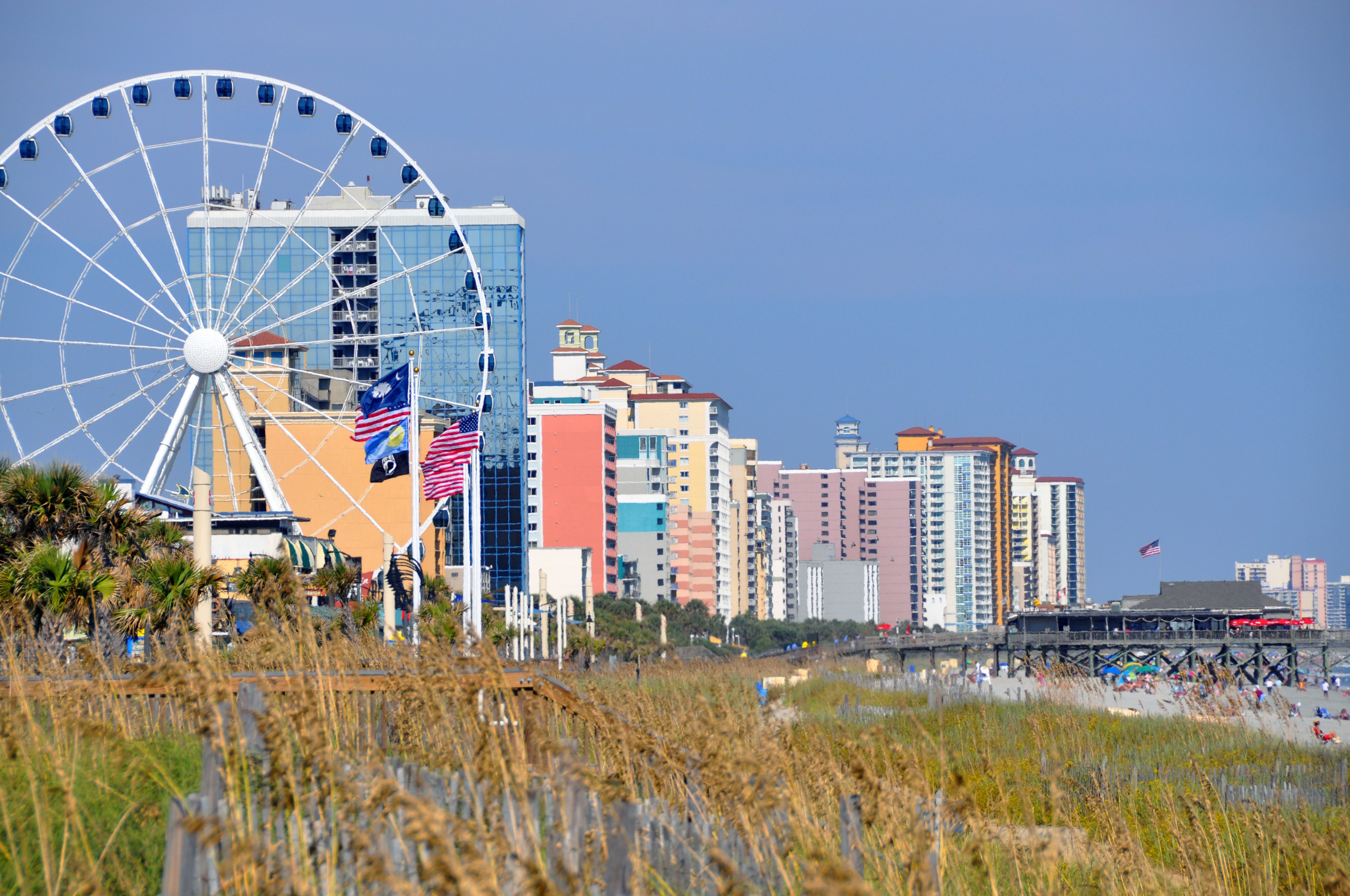 The SkyWheel, oceanfront hotels, and a fishing pier seen from the beach dunes with sea oats; city skyline context along the Boardwalk, not the event.